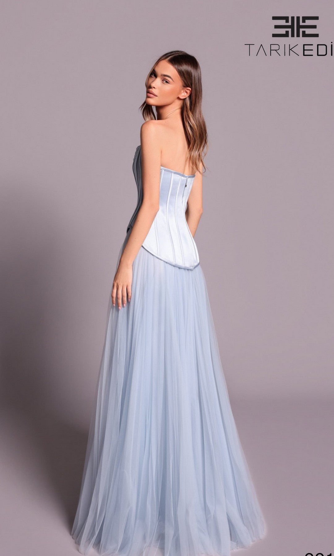 A woman in the Tarik Ediz 99137 long formal dress, featuring a strapless corset bodice and light blue tulle skirt, stands slightly turned away against a plain background.